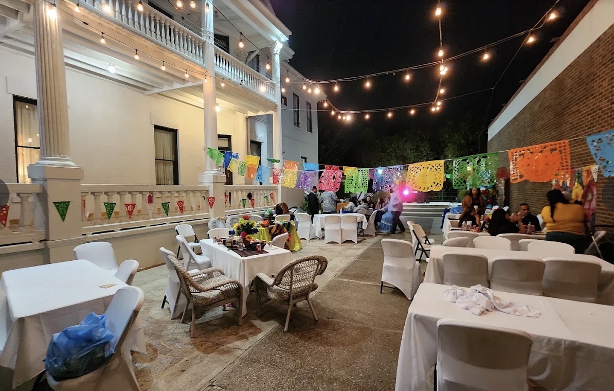 An evening gathering in the mansion's courtyard, with string lights, white-dressed tables and festive banners