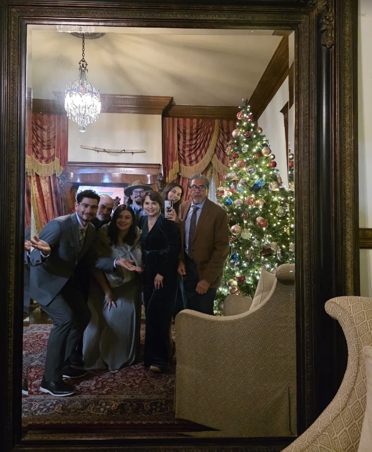 Guests gathered in the mansion's parlor beside a glowing Christmas tree, reflected in a tall gilded mirror