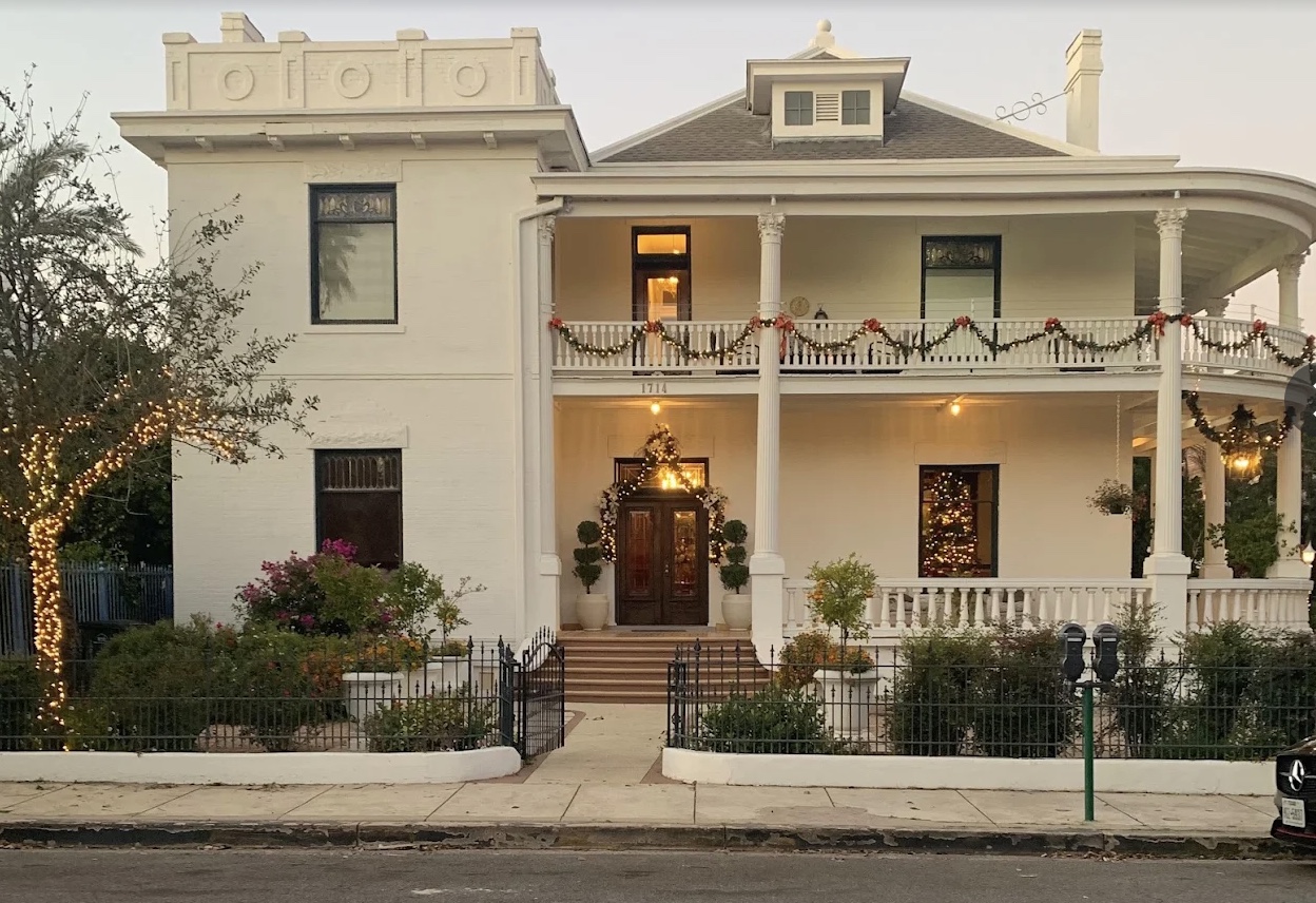 The white historic Laredo Mansion at dusk, its double gallery wrapped in garlands and glowing with warm light