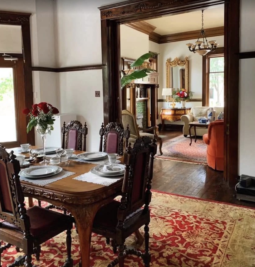 The mansion's dining room with carved mahogany chairs set for tea, opening onto a parlor with gilded mirror and chandelier